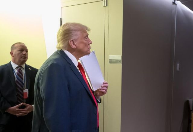 Presidente dos Estados Unidos aparece em pé enquanto aguarda para discursar na 80ª sessão da Assembleia Geral da ONU. Ele assiste a tv, que mostra o presidente do Brasil discursando