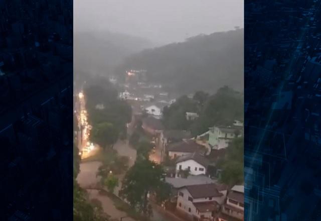 Região montanhosa, com casas à esquerda e área de mata à direita, vista do alto, sob forte chuva e céu cinza e carregado