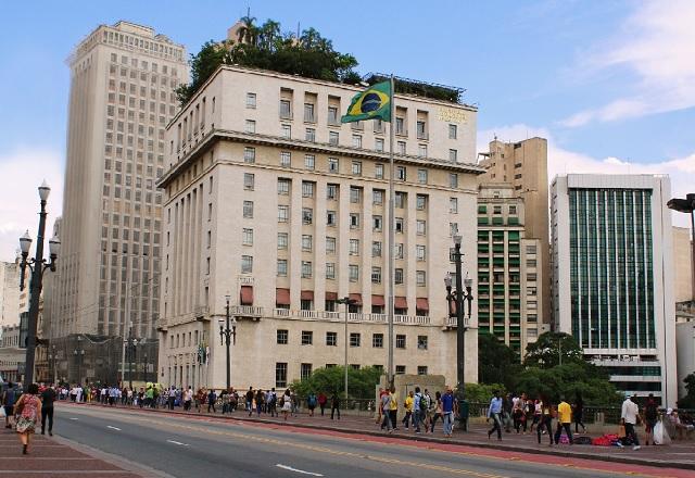 Pessoas passam em frente ao Edifício Matarazzo, próximo do qual há uma bandeira do Brasil hasteada (Guilherme Cunha/SMTUR)