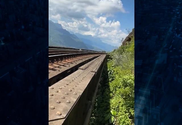 Trilhos de trem em região montanhosa, com sol, céu azul e nuvens