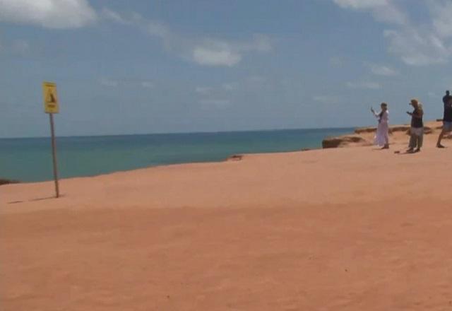 Turistas no alto de falésia, em praia, com mar ao fundo e céu azul, ensolarado