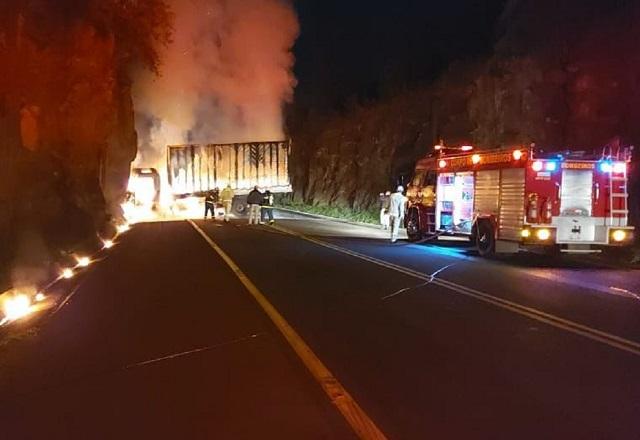 Veículo incendiado bloqueando estrada durante ataque, com carro do corpo de bombeiros ao lado, durante a noite