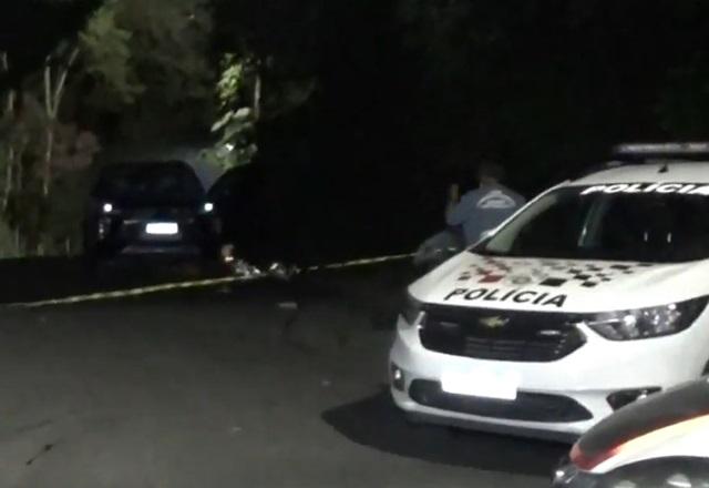 Viatura policial à frente e, ao fundo, corpo de pessoa morta coberta com manta térmica, atrás de faixa de isolamento, durante a noite