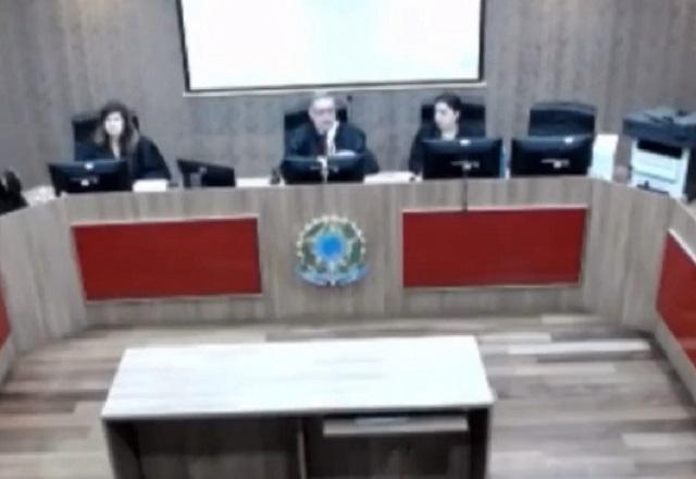 Visão aérea de sala de julgamento, com um juiz no meio do púlpito, ao lado de outros dois profissionais do direito