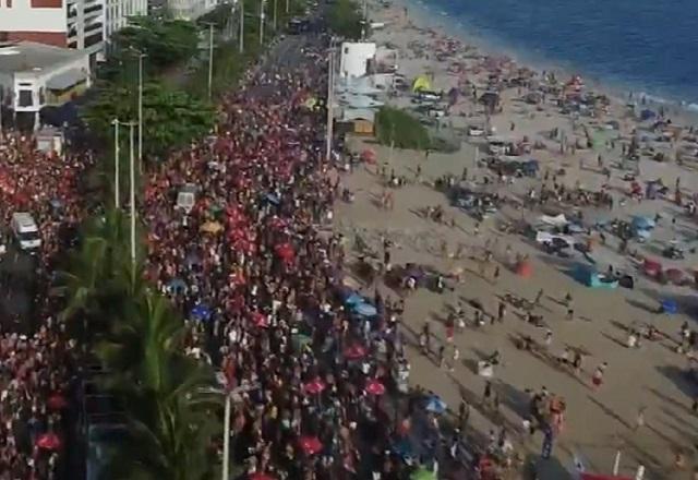 Vista aérea de multidão em praia, durante o dia, com parte da orla e do mar aparecendo
