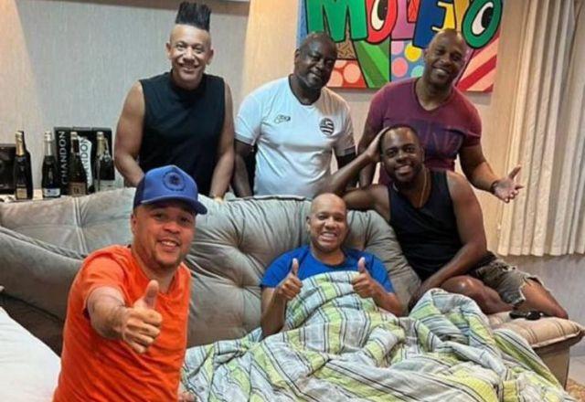 anderson sentado no sofá e cercado dos outros cinco integrantes do grupo Molejo