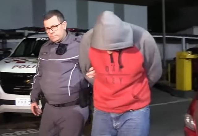 homem de cabeça baixa, blusa de frio vermelha e cinza, algemado com policial ao lado