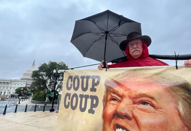 homem de idade, segura guarda chuva com uma faixa com a foto de trump e frase golpe, golpe