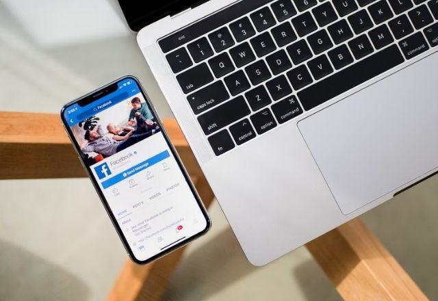 imagem mostra celular com aplicativo facebook e um notebook sobre uma mesa