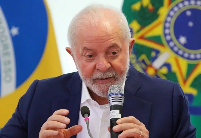 Lula fala ao microfone, próximo à bandeira do Brasil (Agência Brasil)