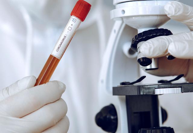 uma pessoa com equipamento de proteção individual manuseia um tubo de vidro escrito coronavírus e analisa um material no microscópio