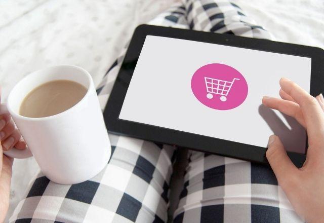 uma pessoa fazendo compras na internet com uma caneca de leite na mão esquerda e na outra segurando um tablet para acessar uma loja virtual